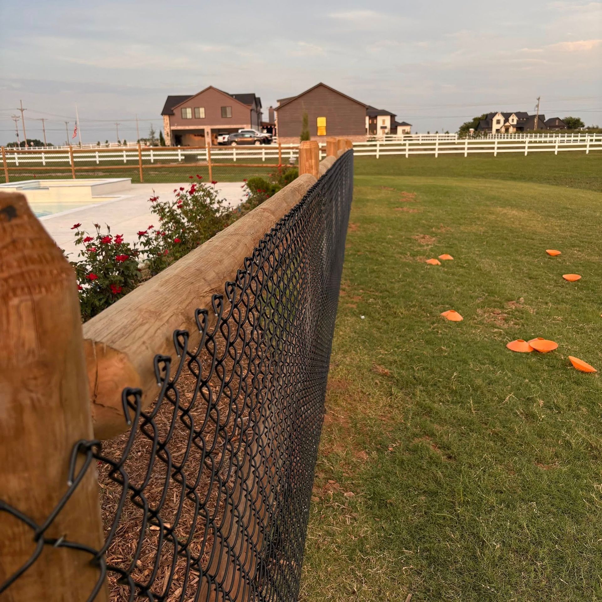 Chain-link fence beside a grassy yard with orange markers, white rails, and houses in the background