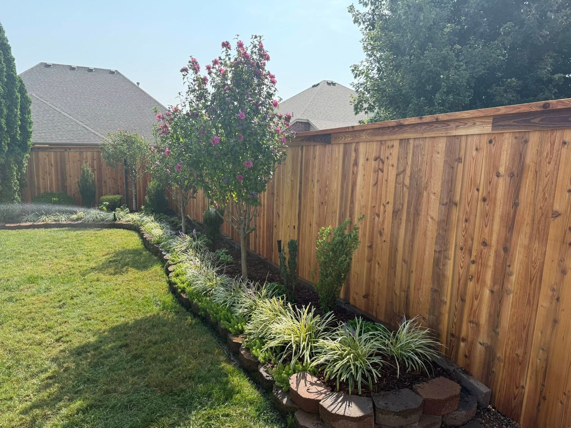 Sunny backyard with a wooden fence, green lawn, and flower bed with shrubs and potted plants.