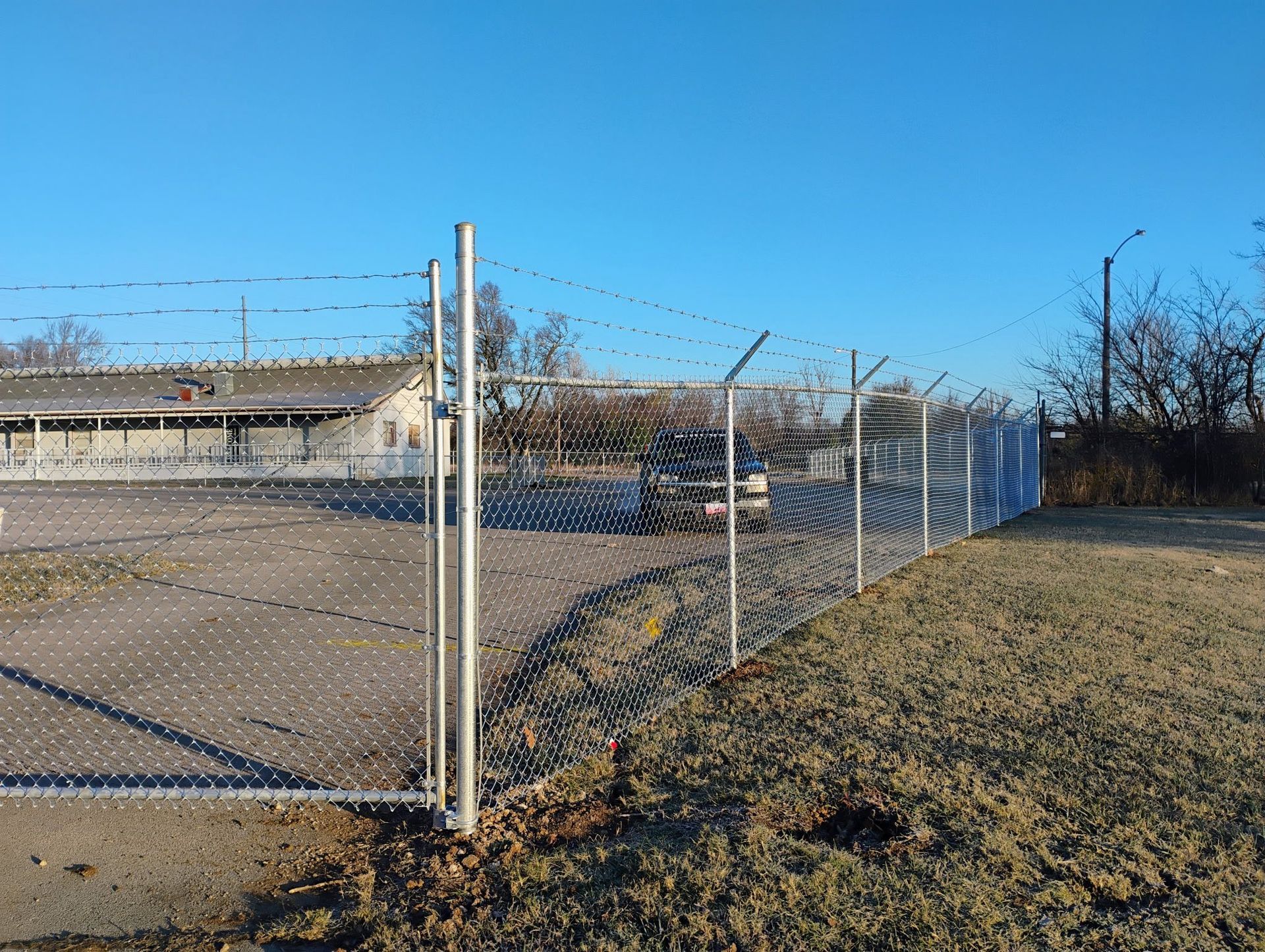 Chain-link fence along a paved lot beside dry grass under a clear blue sky.