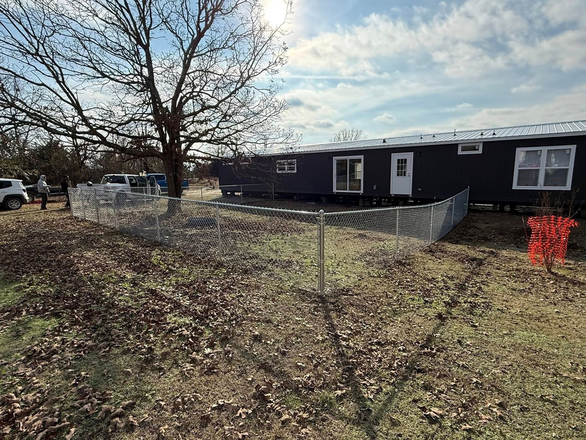 Leaf-covered yard beside a black mobile home under a cloudy sky, with a tree and chain-link fence