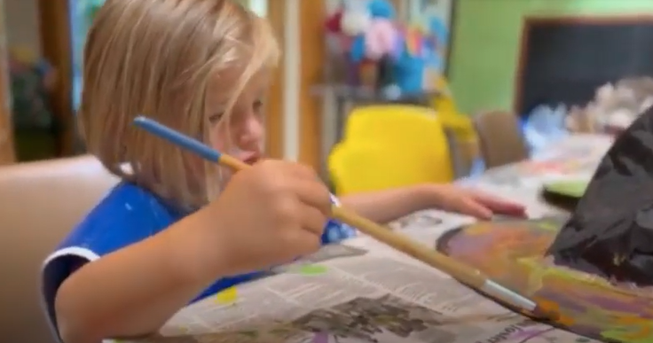 A little girl is sitting at a table painting with a brush