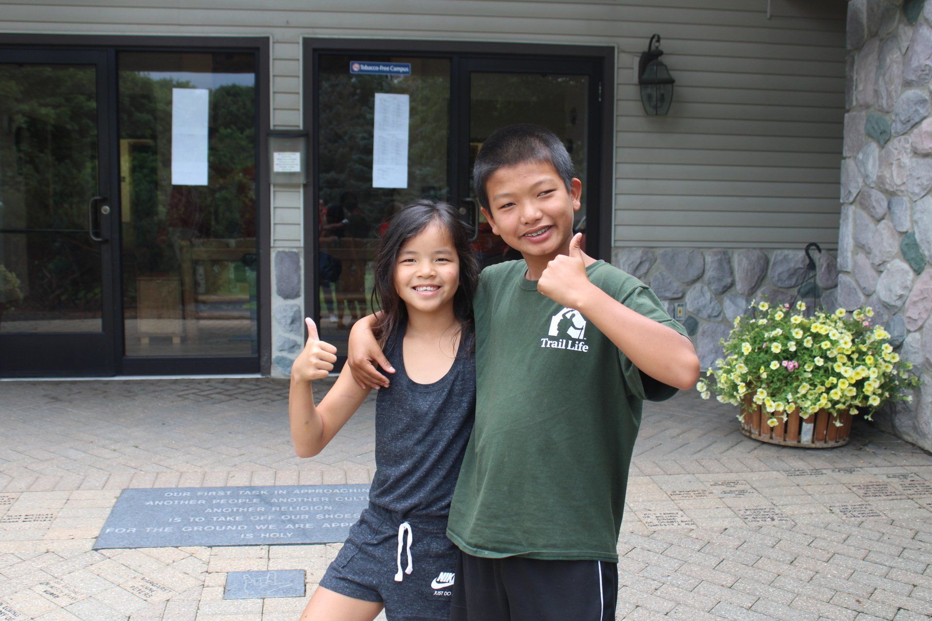 A boy and a girl are giving a thumbs up in front of a building.