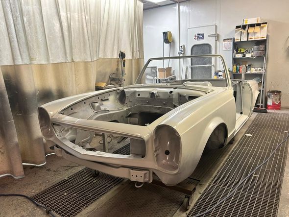 Un operaio, coperto di polvere e con indosso indumenti protettivi, sta sabbiando la carrozzeria rossa di un'auto in un'officina di verniciatura.
