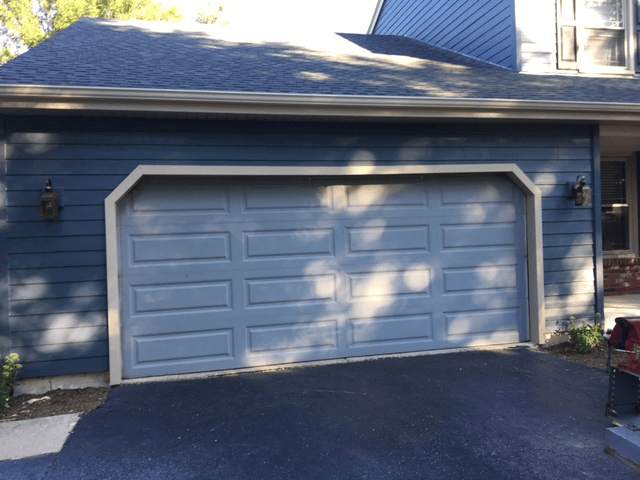 Blue house garage with white double doors and black asphalt driveway, dappled in sunlight