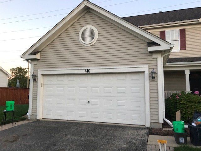 Detached garage with white double doors and beige siding next to a house, with a driveway in front.