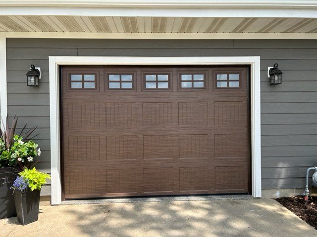 Brown garage door with four small windows, framed by white trim, on a gray house exterior