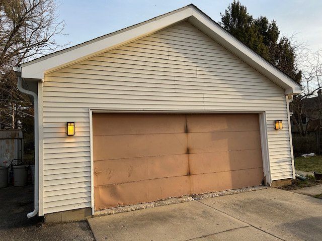 Detached white garage with a brown door and lit wall lamps beside a driveway