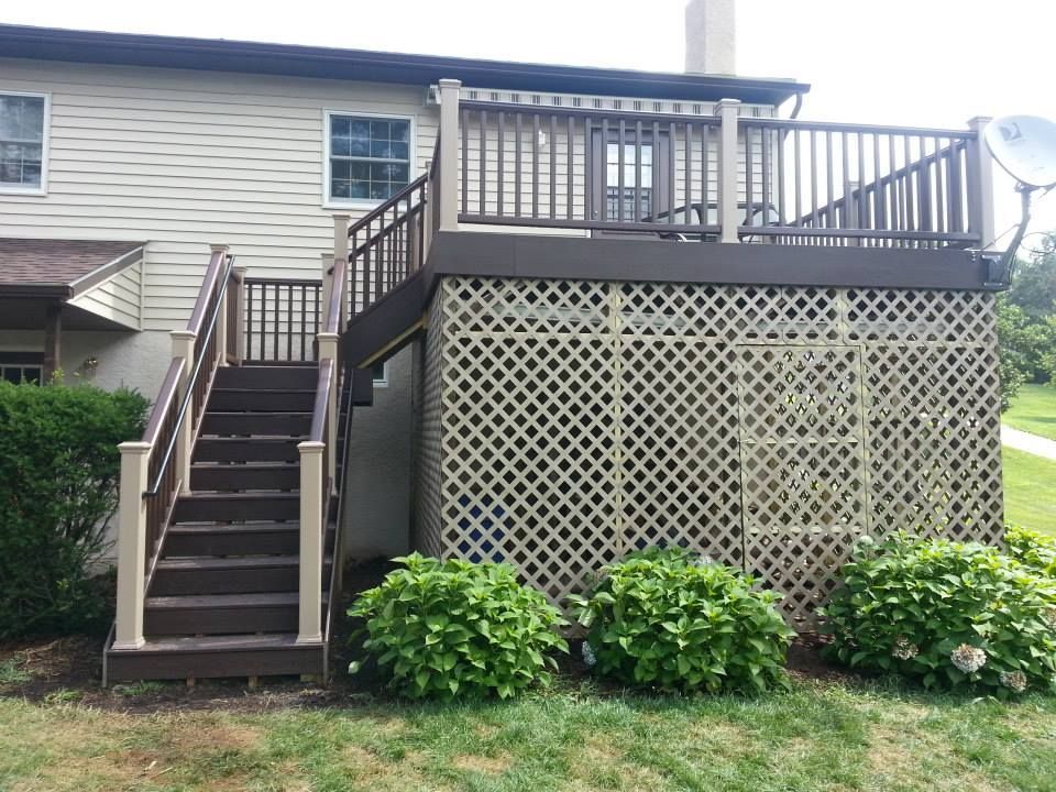 Two-story deck with stairs, latticework, and satellite dish attached to a beige house, surrounded by green shrubs.