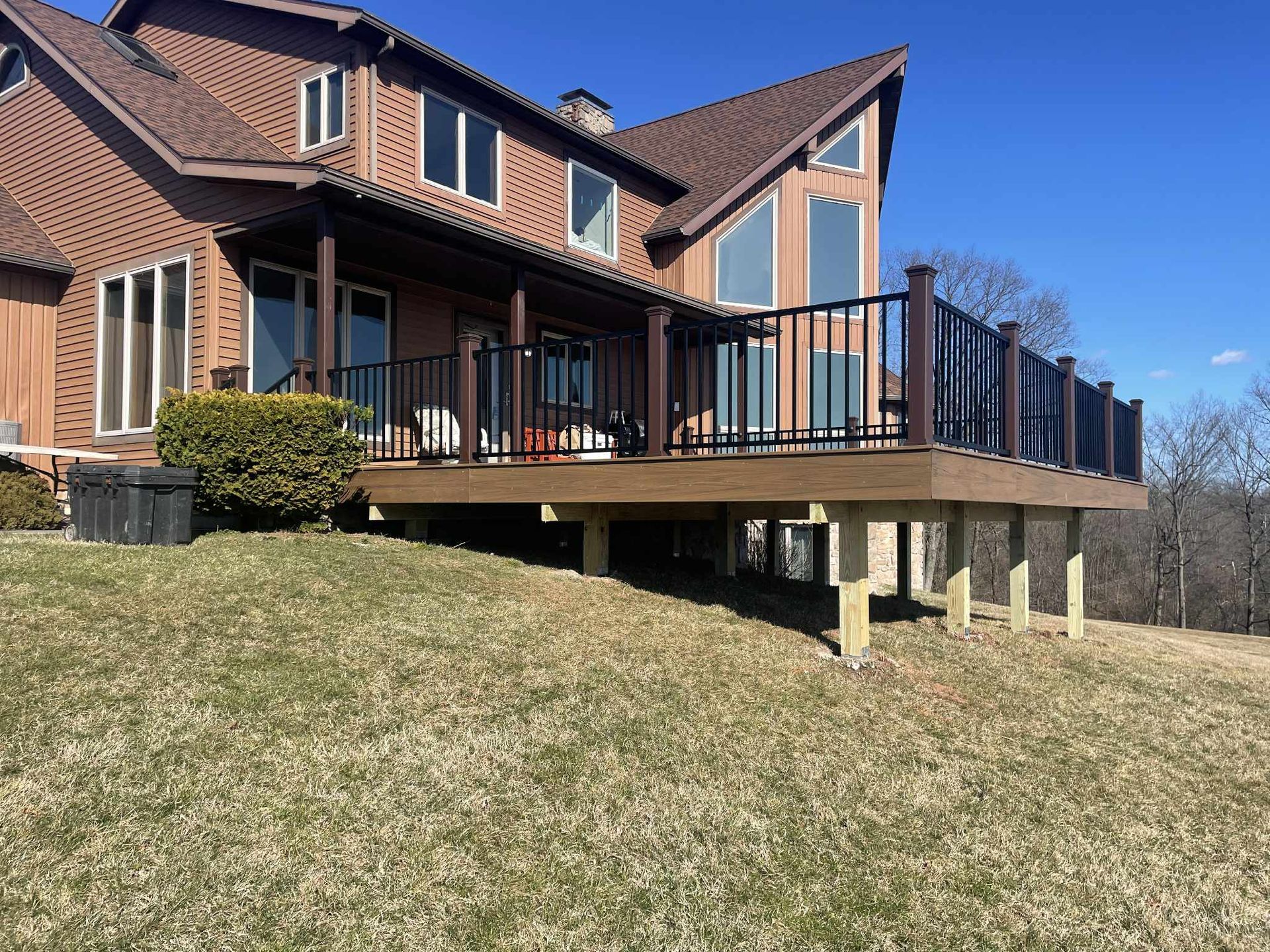 Brown house with a large deck, supported by wooden posts, overlooking a grassy hill on a sunny day.
