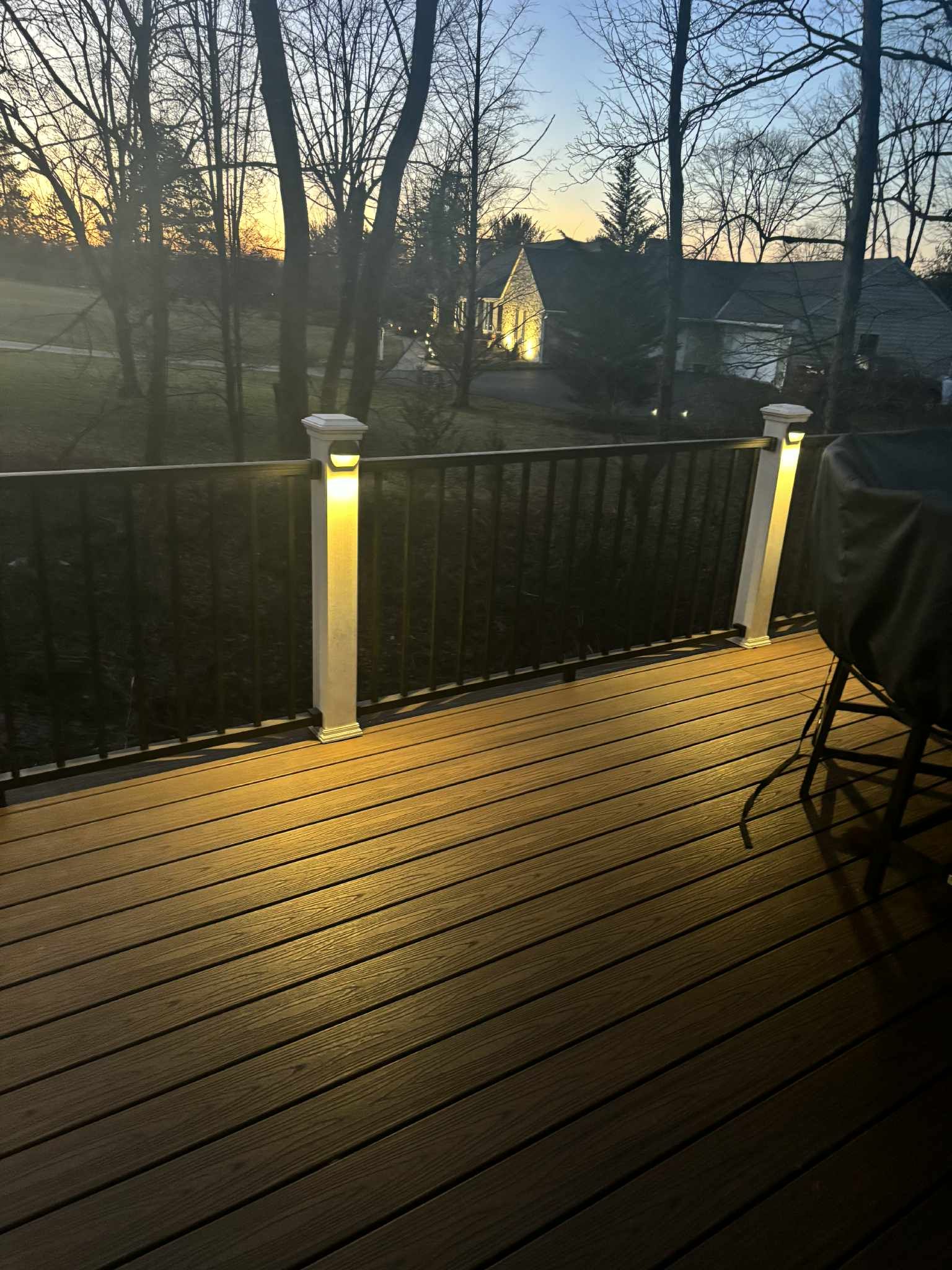Deck with illuminated posts at dusk, overlooking a yard and trees.