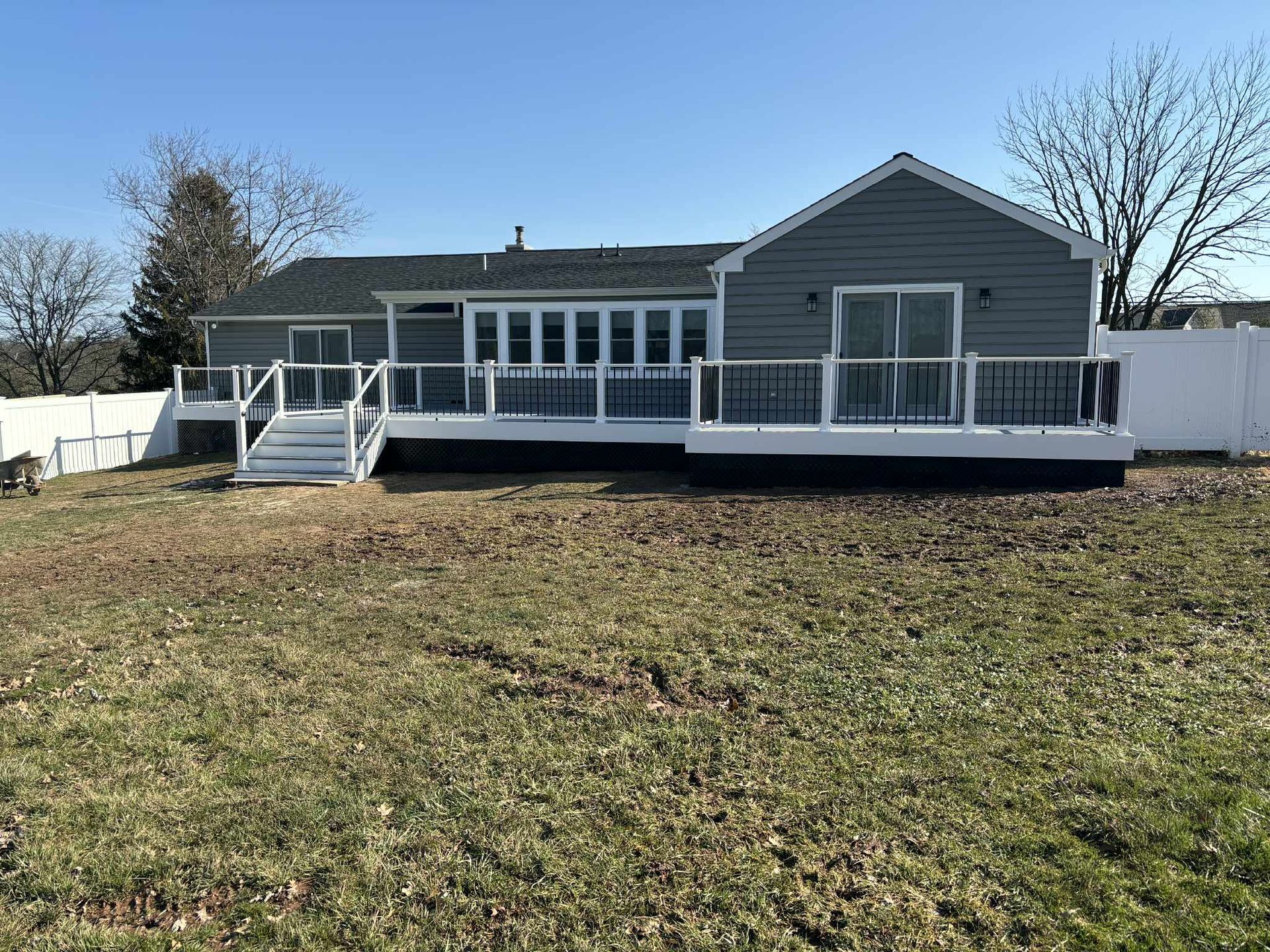 Back view of a gray house with a large deck, white railing, and grassy yard on a sunny day.