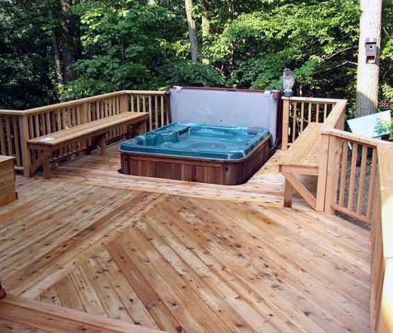 Wooden deck with a hot tub surrounded by benches in a wooded setting.