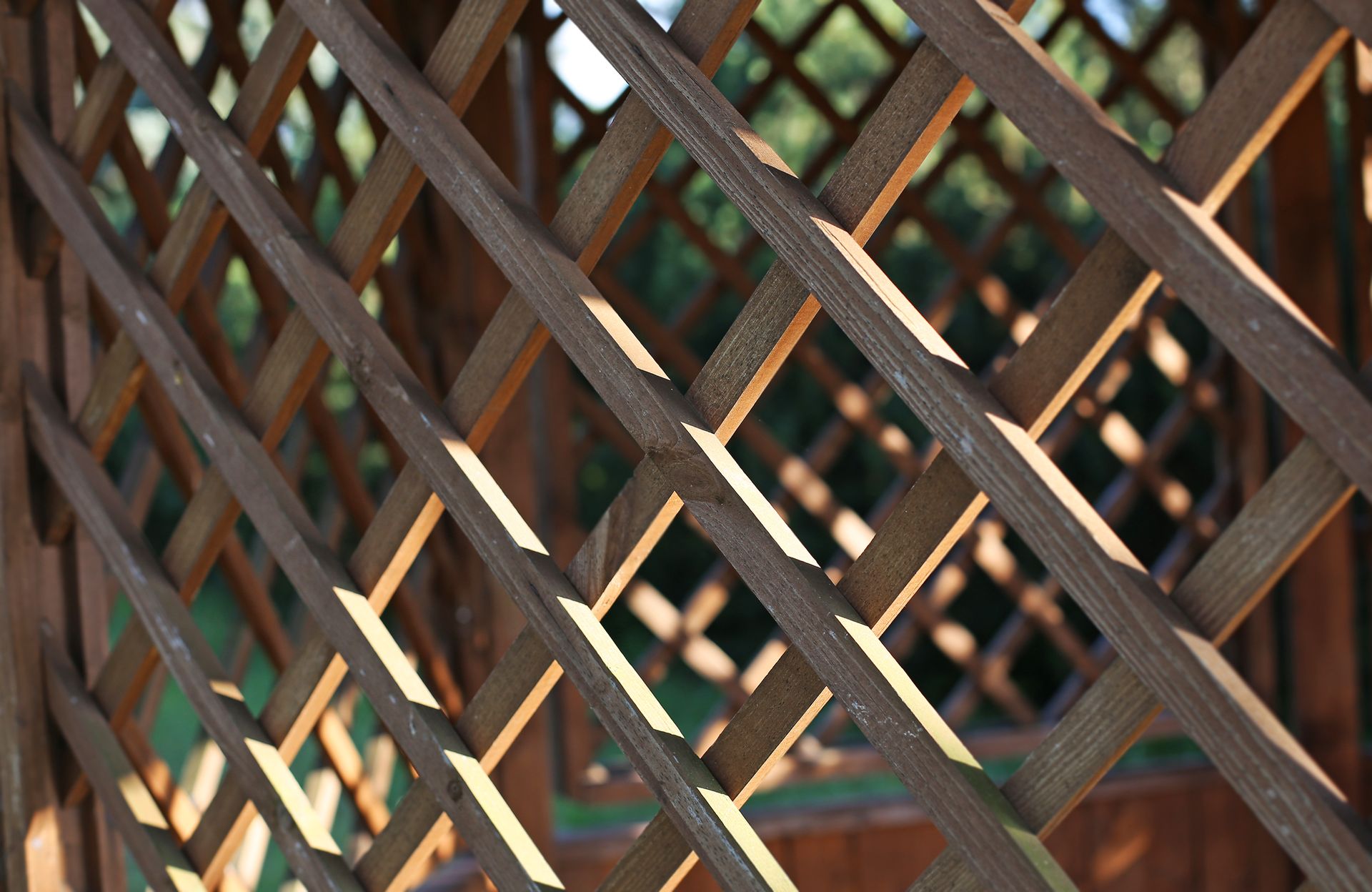 Wooden lattice structure with diamond-shaped openings. Sunlight casts shadows, with a green background.
