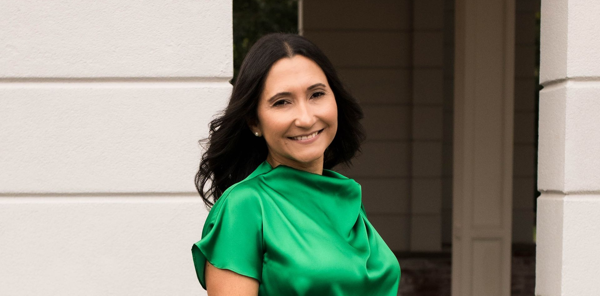 Woman in green top and white skirt smiles outdoors near a building.