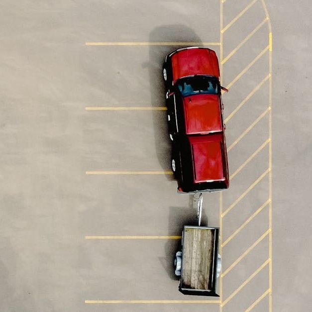 Eine Draufsicht auf einen roten Pickup-Truck, der einen kleinen Anhänger zieht und über mehrere Parkplätze geparkt ist.