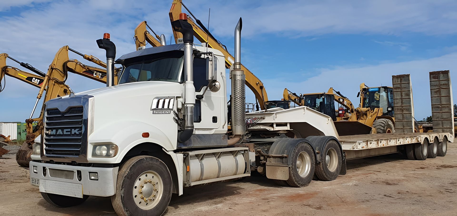 White semi-truck hauling a lowboy trailer loaded with heavy construction equipment at a worksite — Compass Equipment Hire in Mullumbimby, NSW