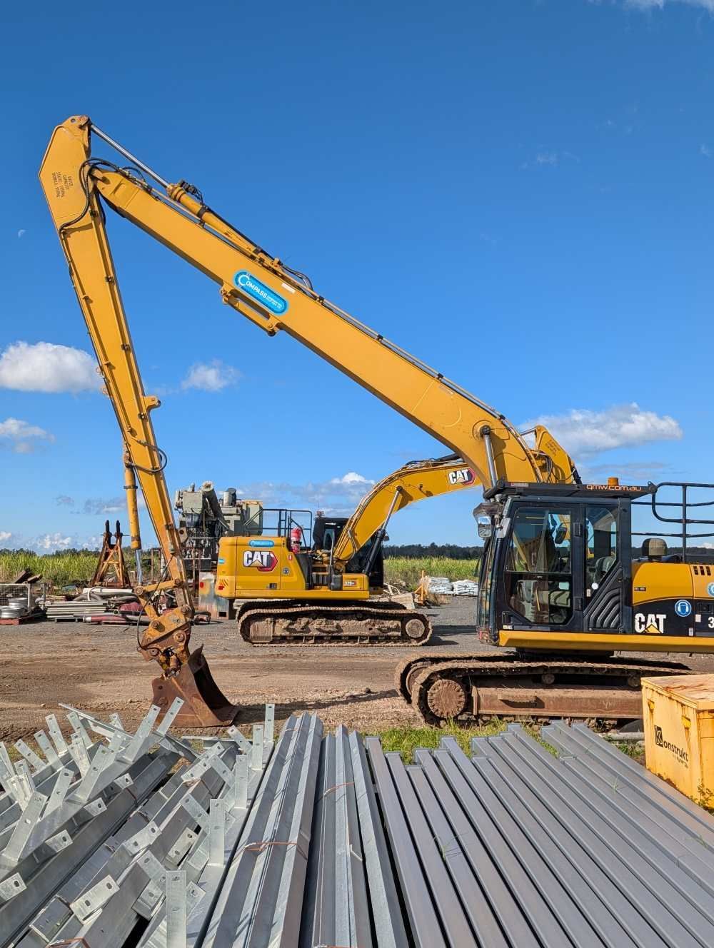 Yellow excavators in a construction yard beside stacked metal beams under a blue sky — Compass Equipment Hire in Byron Bay, NSW