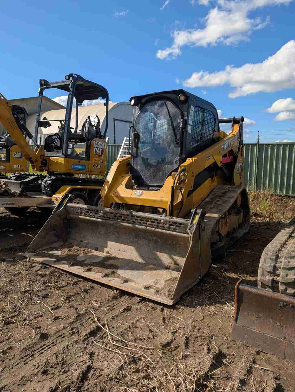 Yellow compact track loader with bucket parked on a muddy lot under a blue sky — Compass Equipment Hire in Mullumbimby, NSW