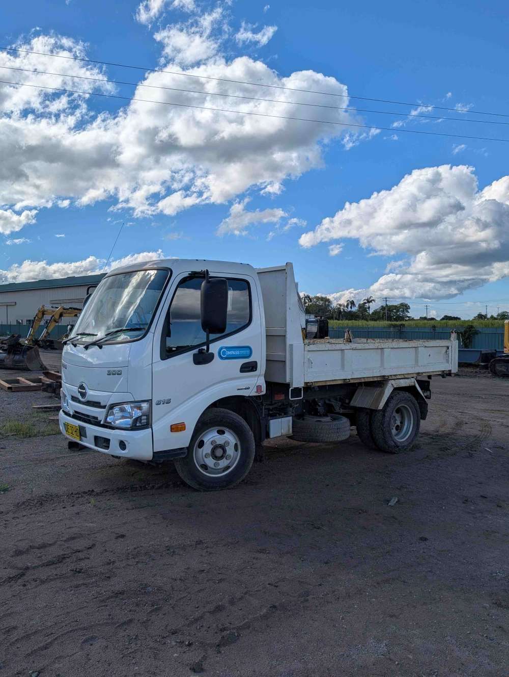 White flatbed truck parked on a gravel lot under a bright blue sky with clouds — Compass Equipment Hire in Kyogle, NSW