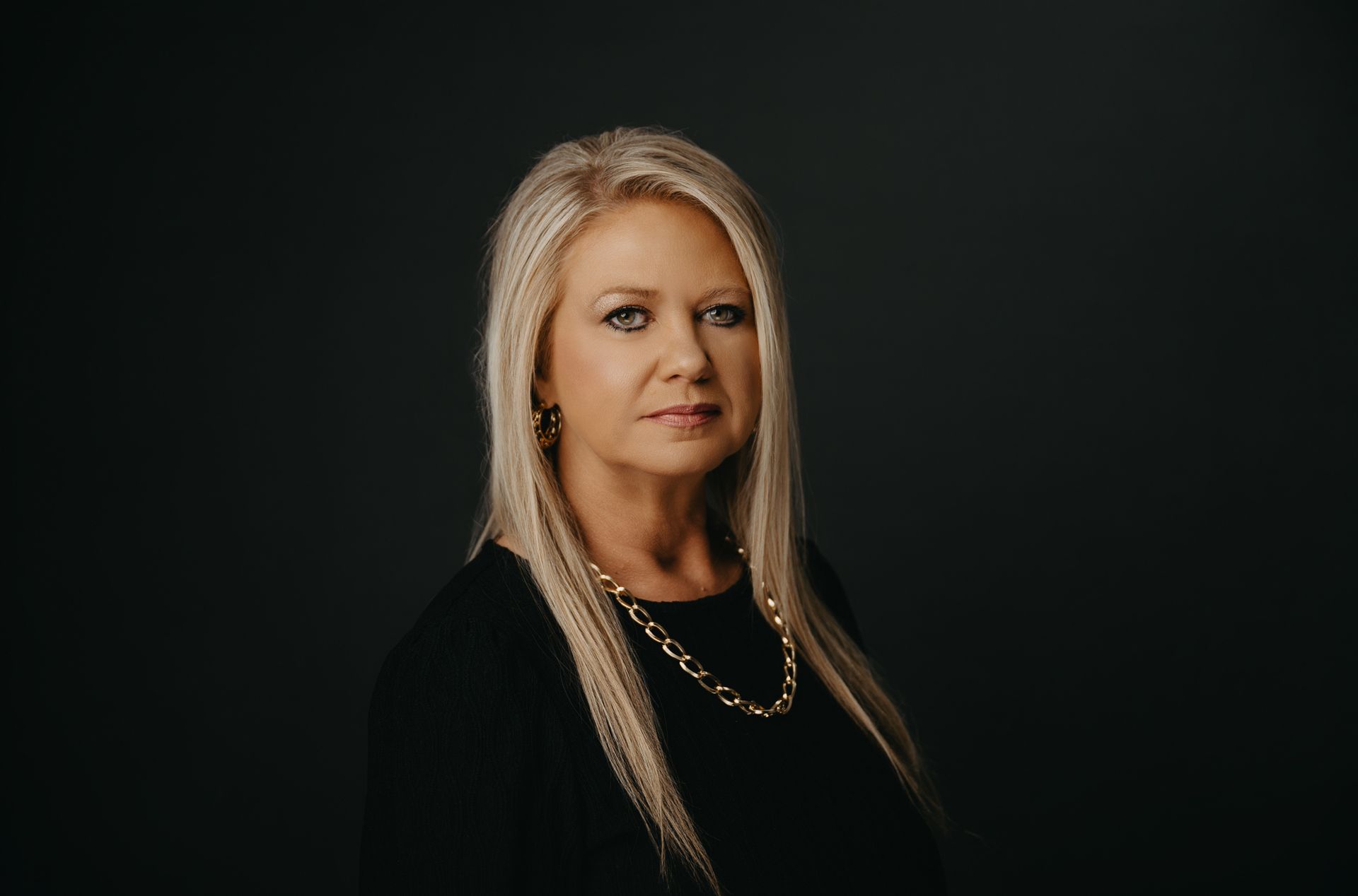 A professional portrait of a person with long blonde hair, wearing a black shirt and gold chain necklace, against black.