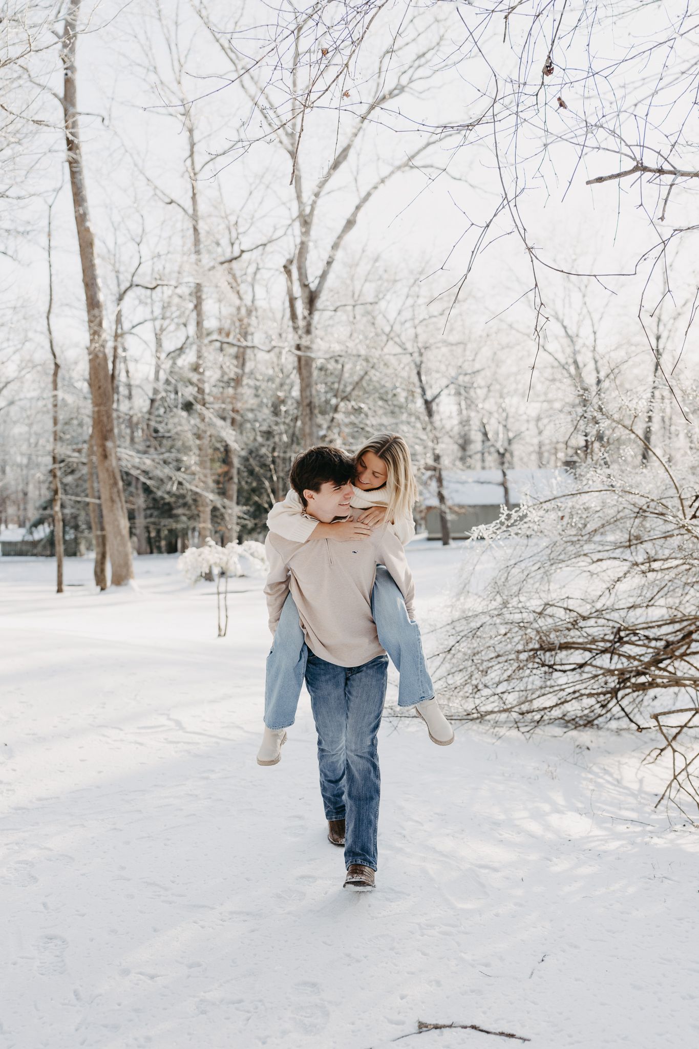 A person gives another person a piggyback ride through a snowy, wooded area, both smiling and wearing winter clothing.