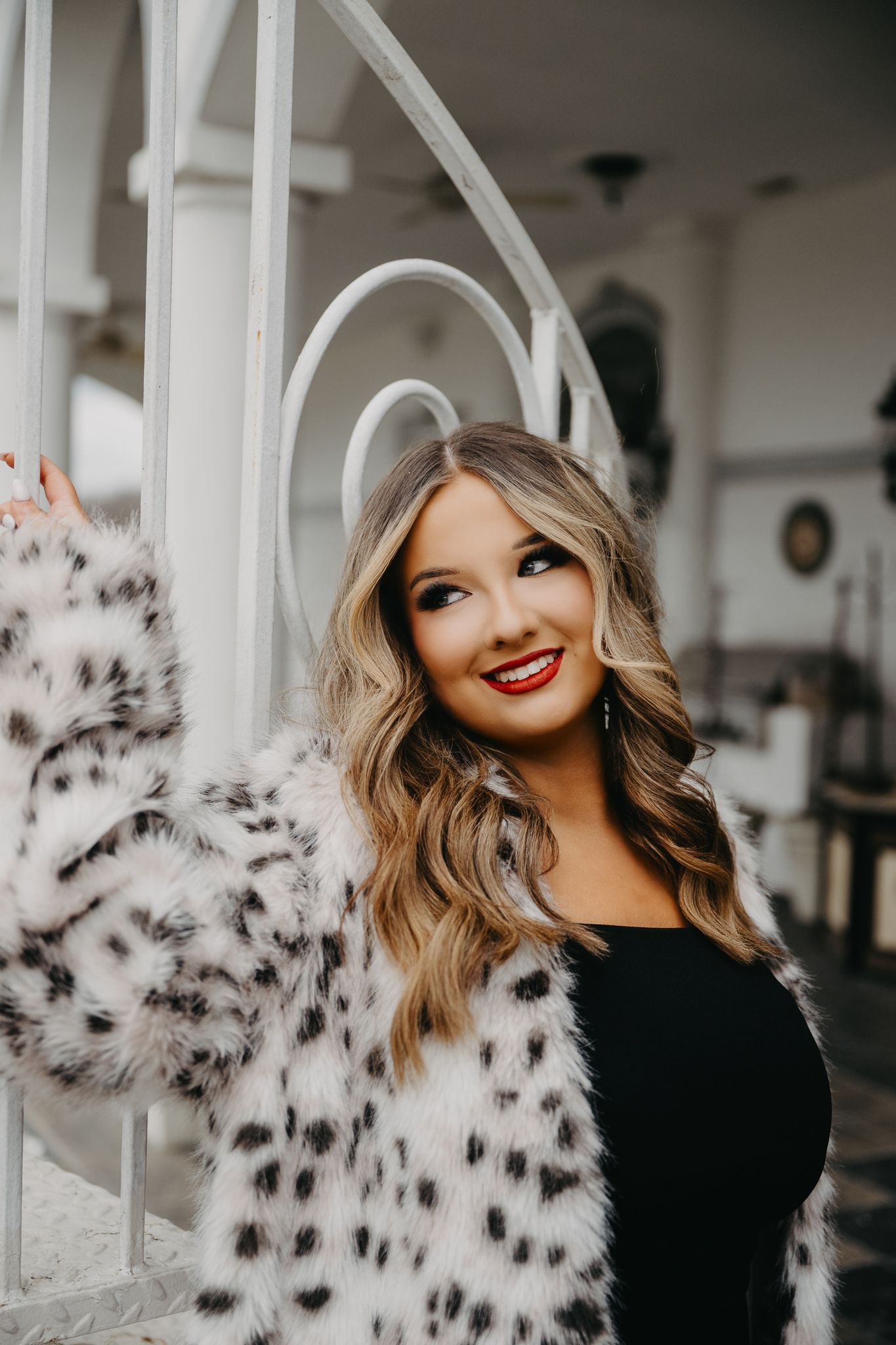 A person with wavy blonde hair wearing a black top and a white, black-spotted faux fur coat leans against a white railing.