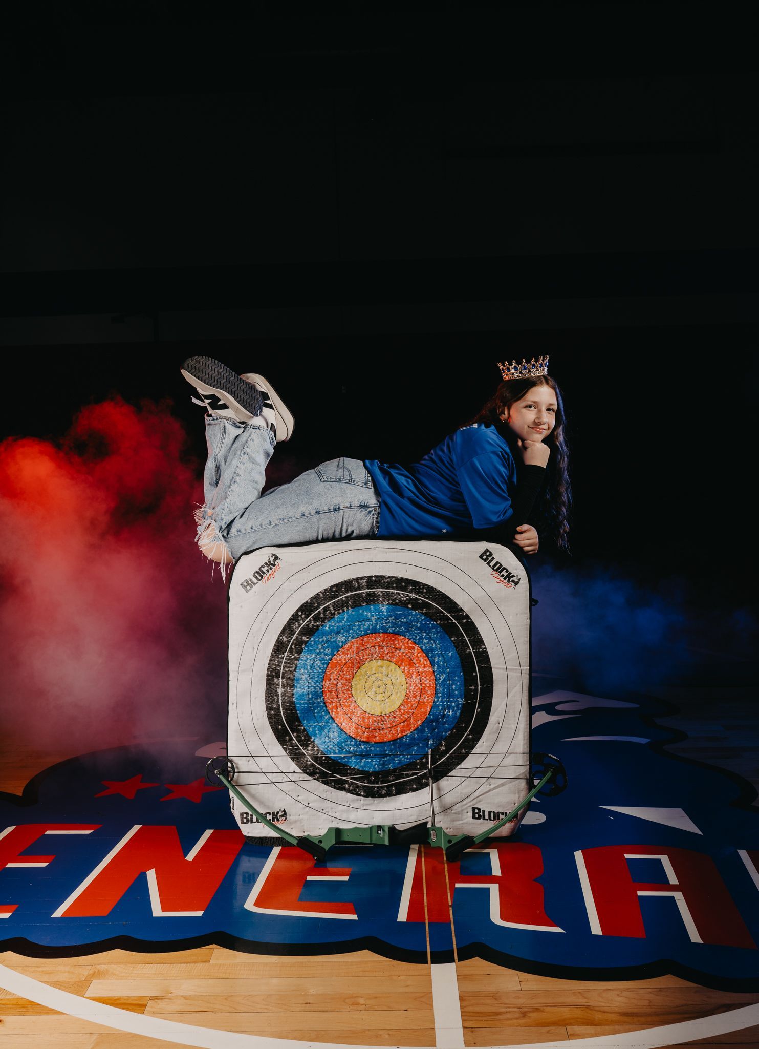 A person lying on an archery target on a gymnasium floor with red and blue smoke effects in the background.