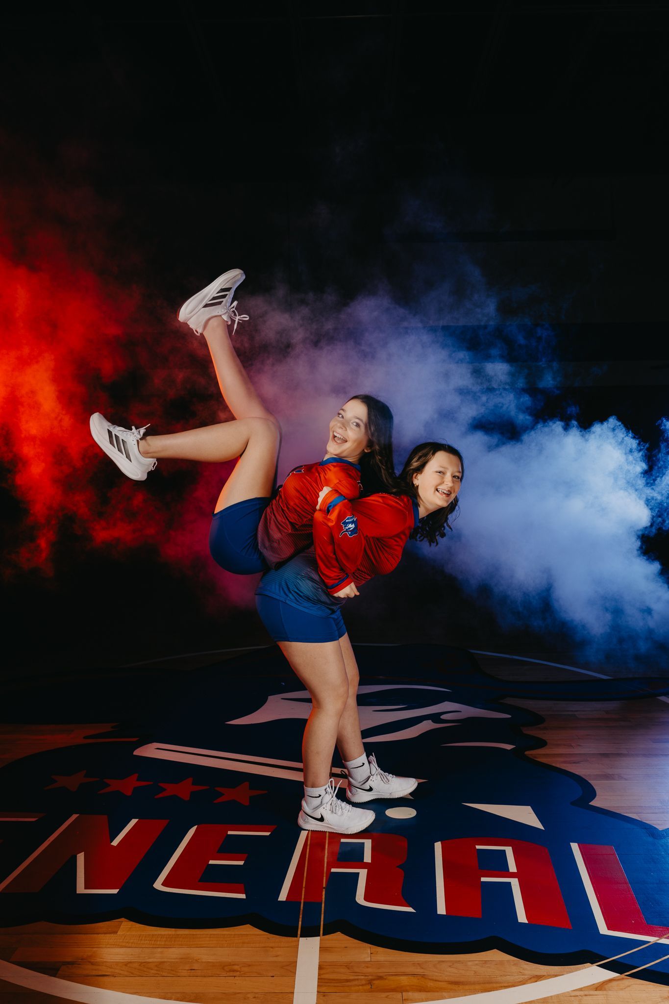 Two people posing back-to-back on a basketball court, surrounded by red and blue smoke, with one person lifted on the back.