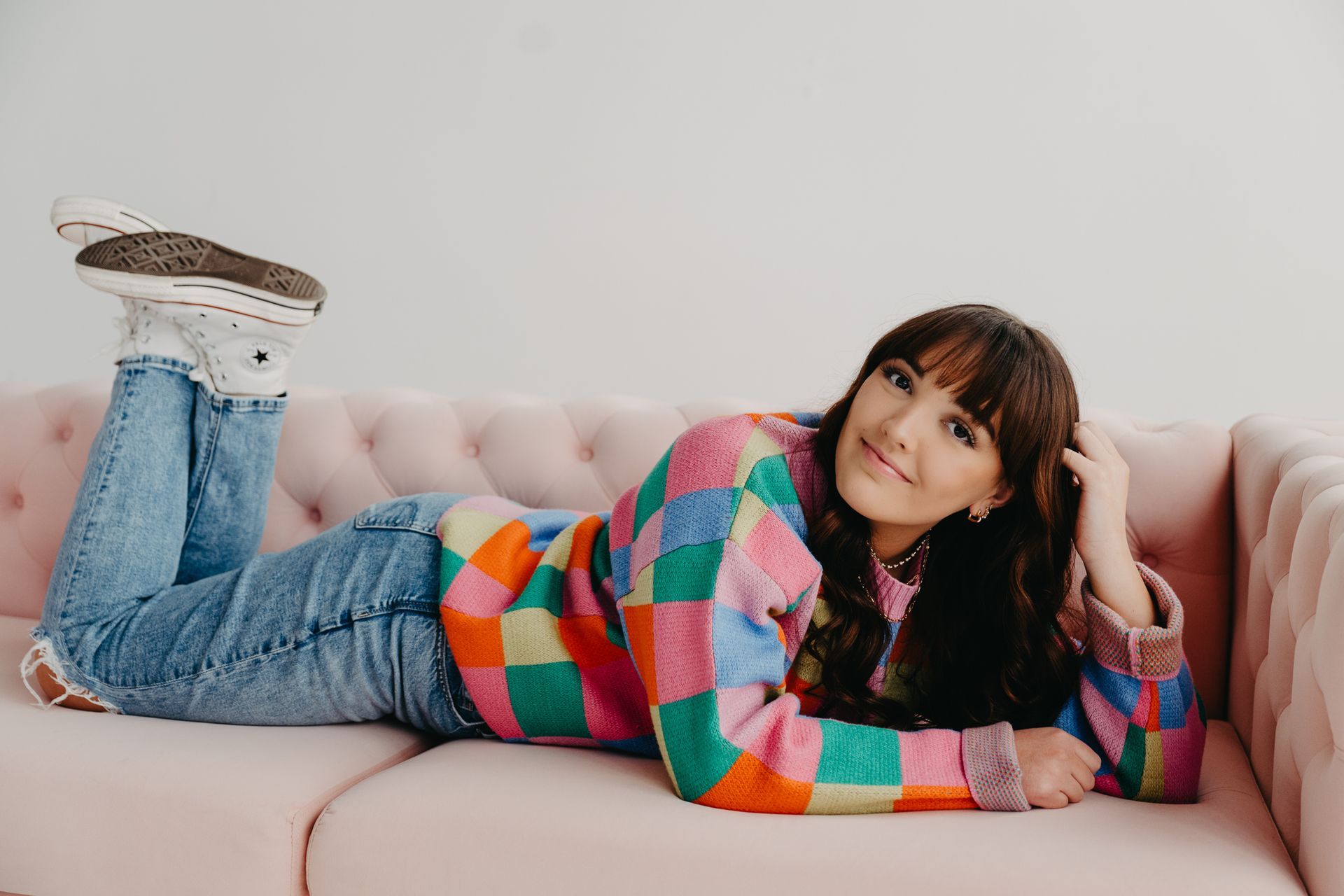 A person with bangs lies on a pink tufted couch, wearing a colorful checkered sweater and jeans, smiling at the camera.