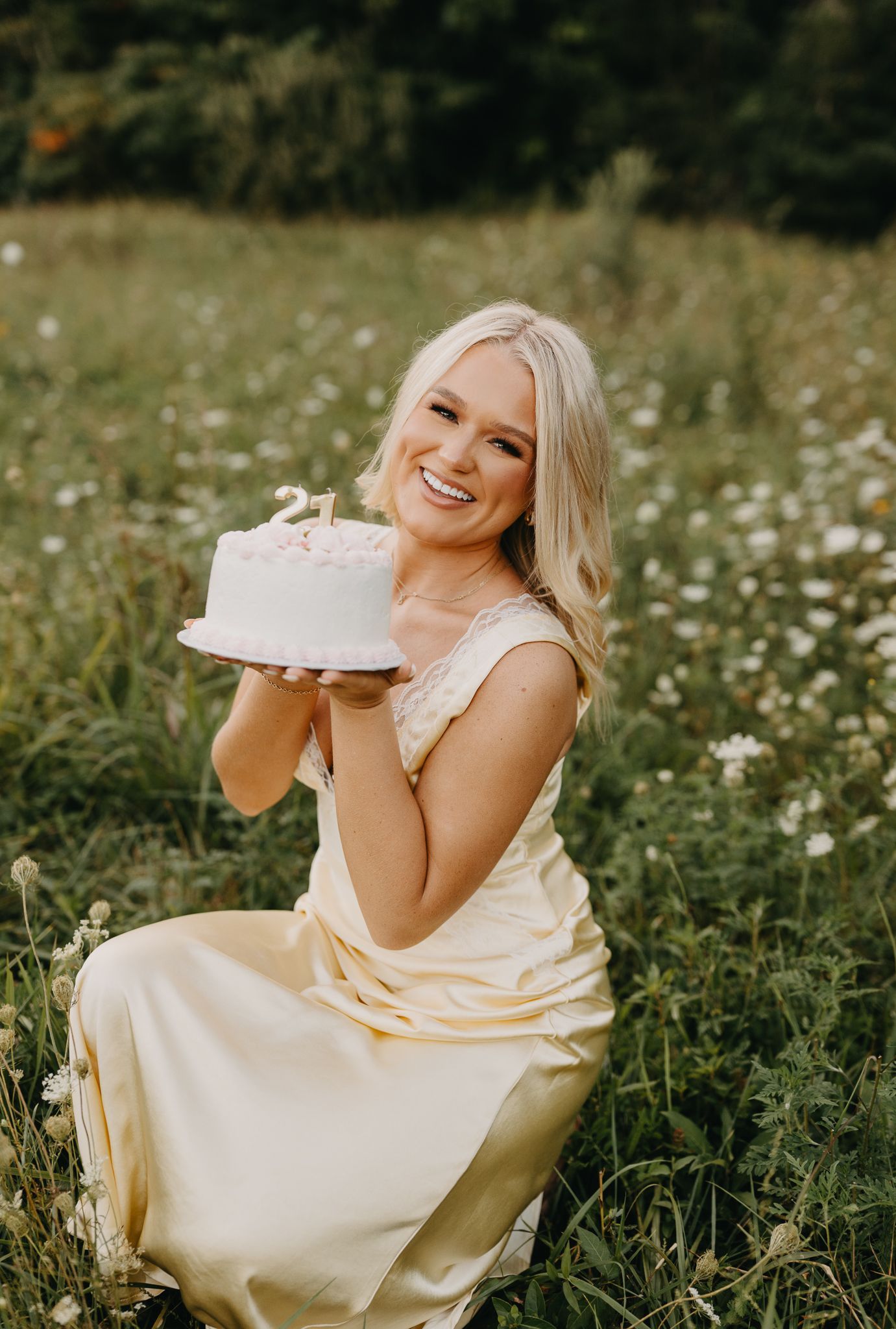 A person in a yellow dress kneeling in a grassy field, smiling while holding a small cake topped with the number 21.