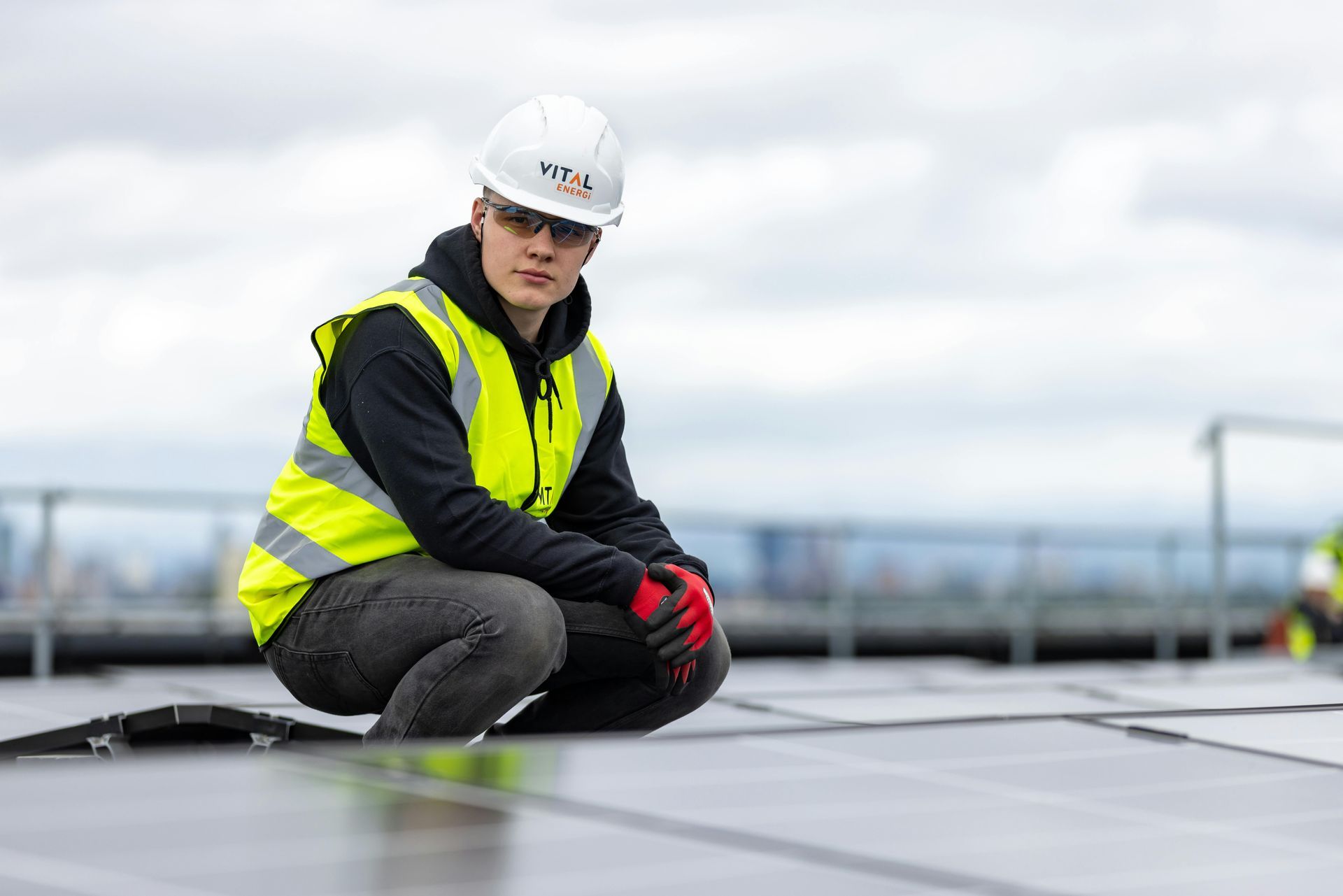 Een werknemer met een veiligheidshelm en -vest knielt op een dak om zonnepanelen te inspecteren.