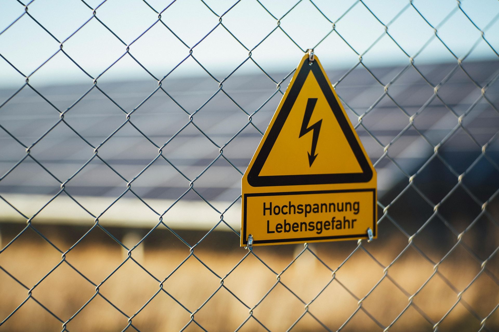 Geel waarschuwingsbord voor hoogspanning op een gaashekwerk met Duitse tekst en bliksemsymbool.