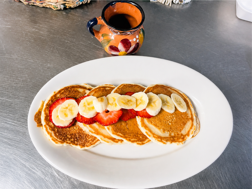 Plate of pancakes topped with banana slices and strawberries, with a small cup of syrup on the table
