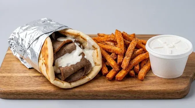Pita wrap with meat and sauce, sweet potato fries, and a cup of dipping sauce on a wooden board