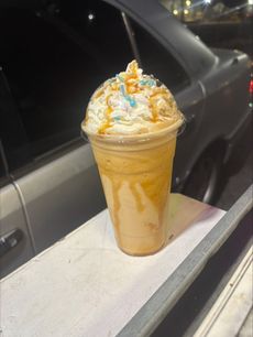 Caramel frappe topped with whipped cream, sprinkles, and caramel drizzle, in a clear plastic cup.