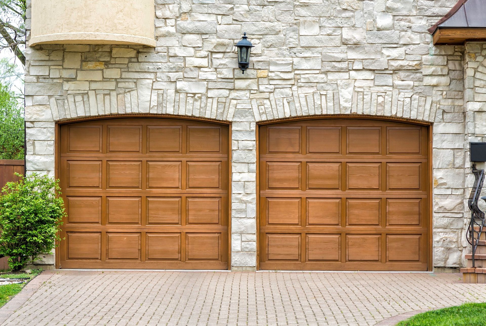 Two wooden garage doors on a stone house facade with a paved driveway and small wall lanterns.