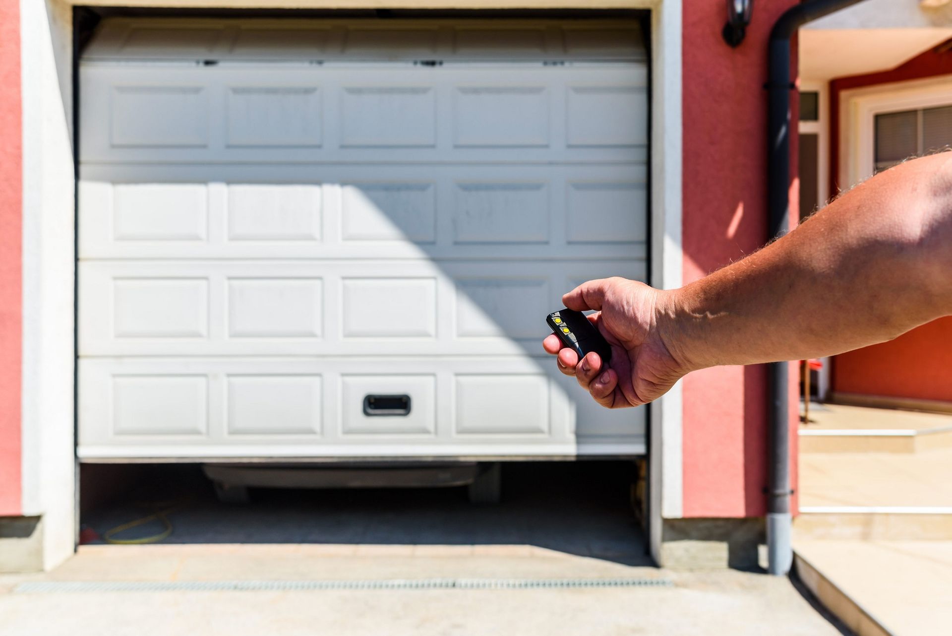Hand holding a garage door opener toward a white garage door in a sunlit driveway