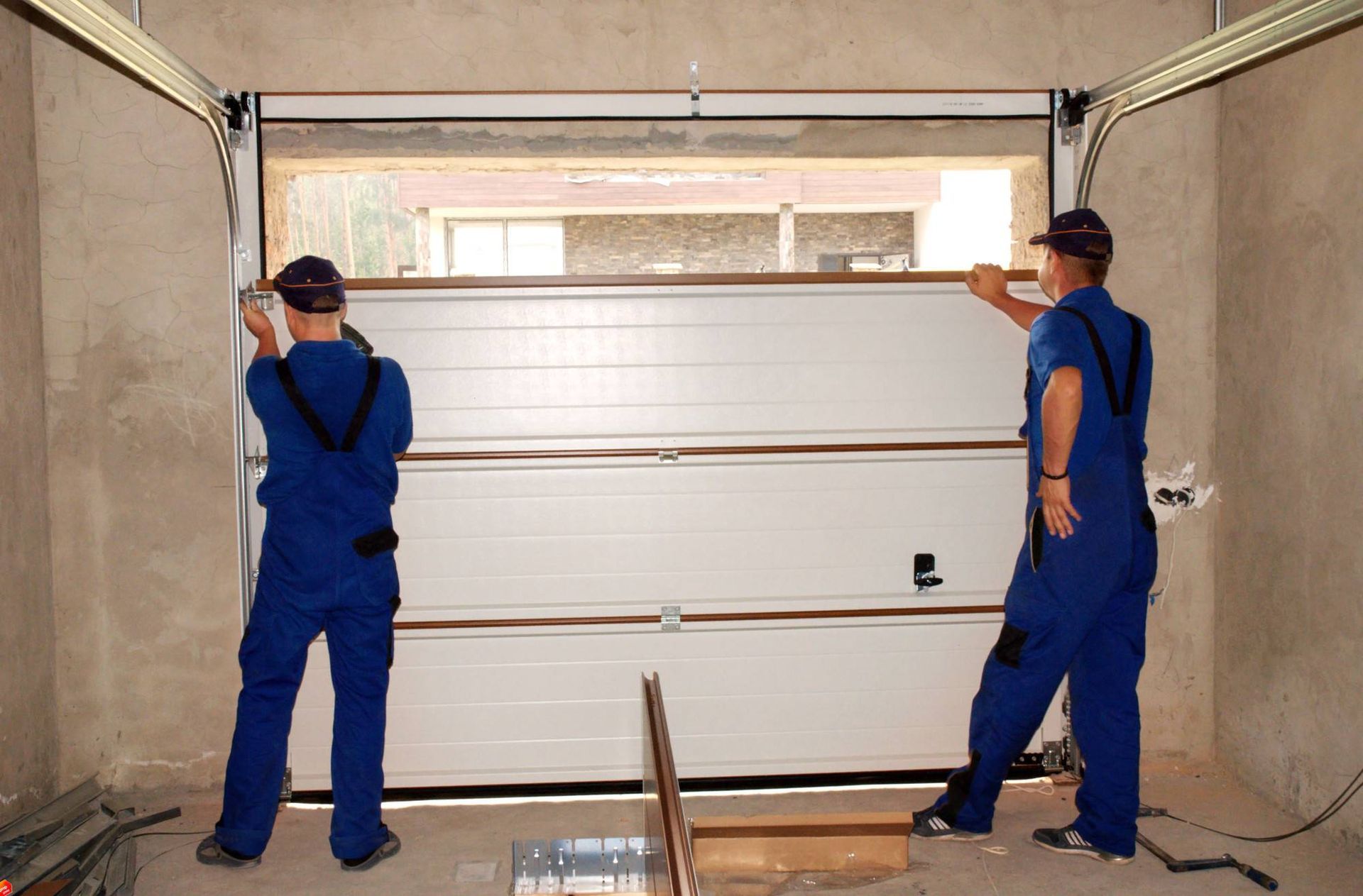 Two workers in blue overalls installing a white garage door inside a building frame