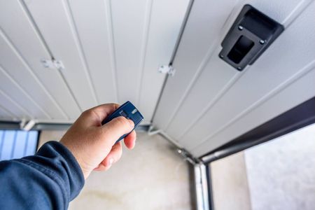 Hand holding blue key fob near a black garage door opener button under a white ceiling