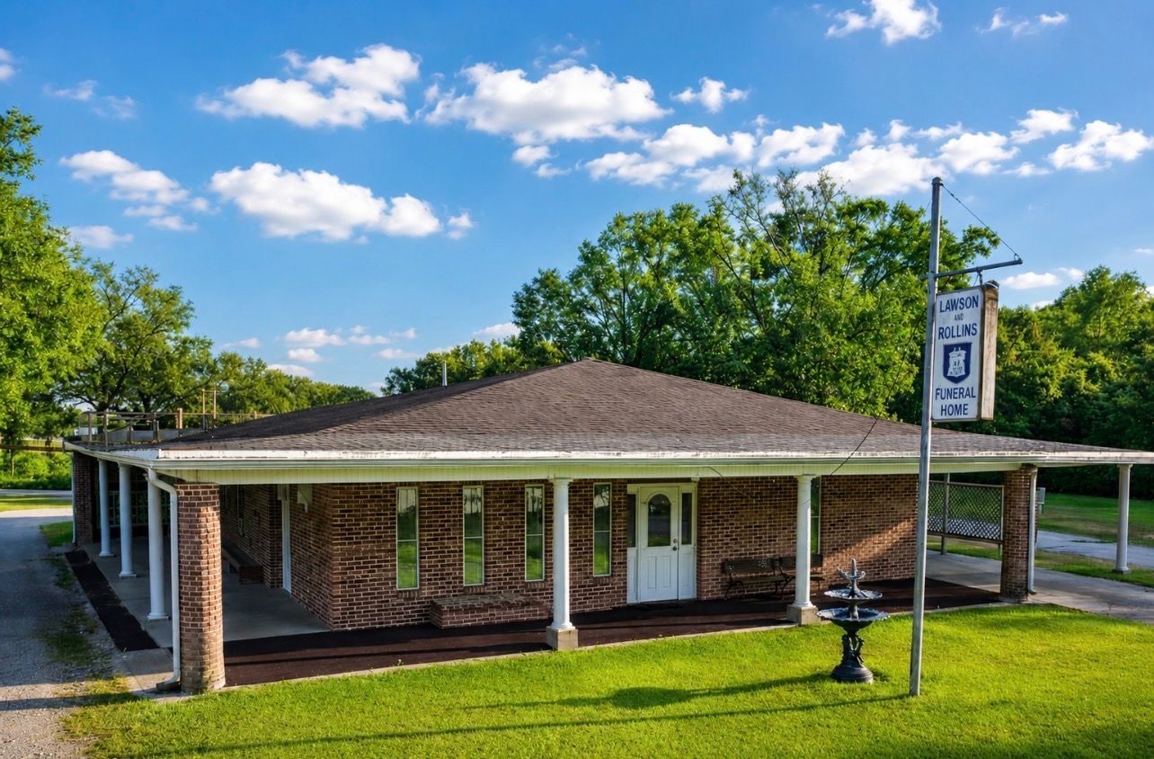 Brick roadside building with a covered carport, parked van, and grassy lawn under trees
