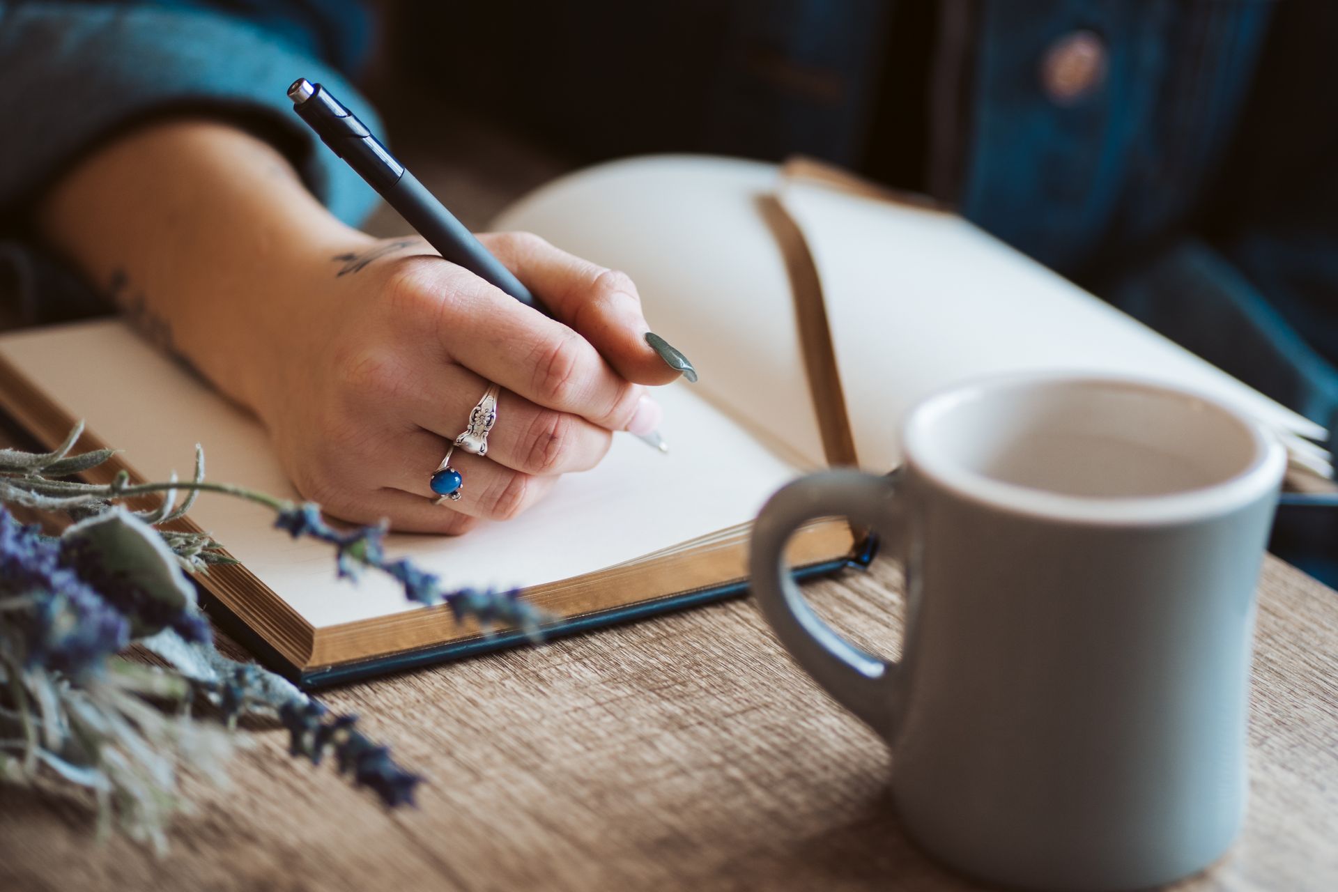 Hand writing in a notebook beside a white mug on a rustic wooden table