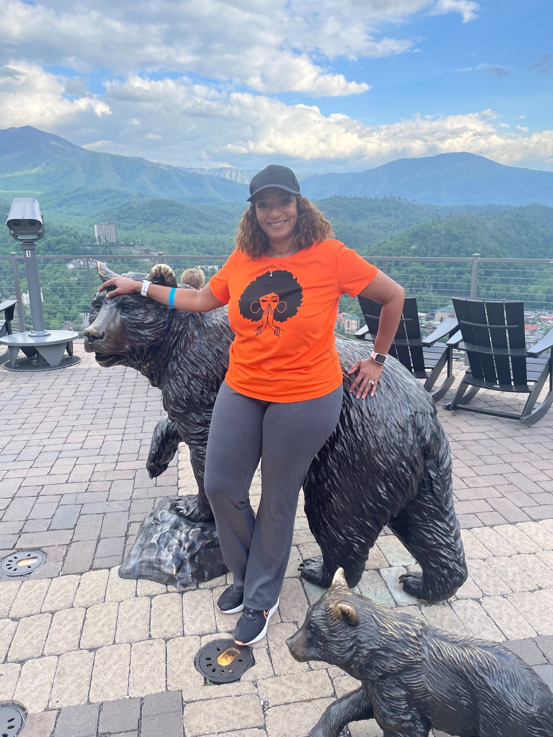 Woman in orange shirt standing with bear statues on a mountain overlook with scenic hills.