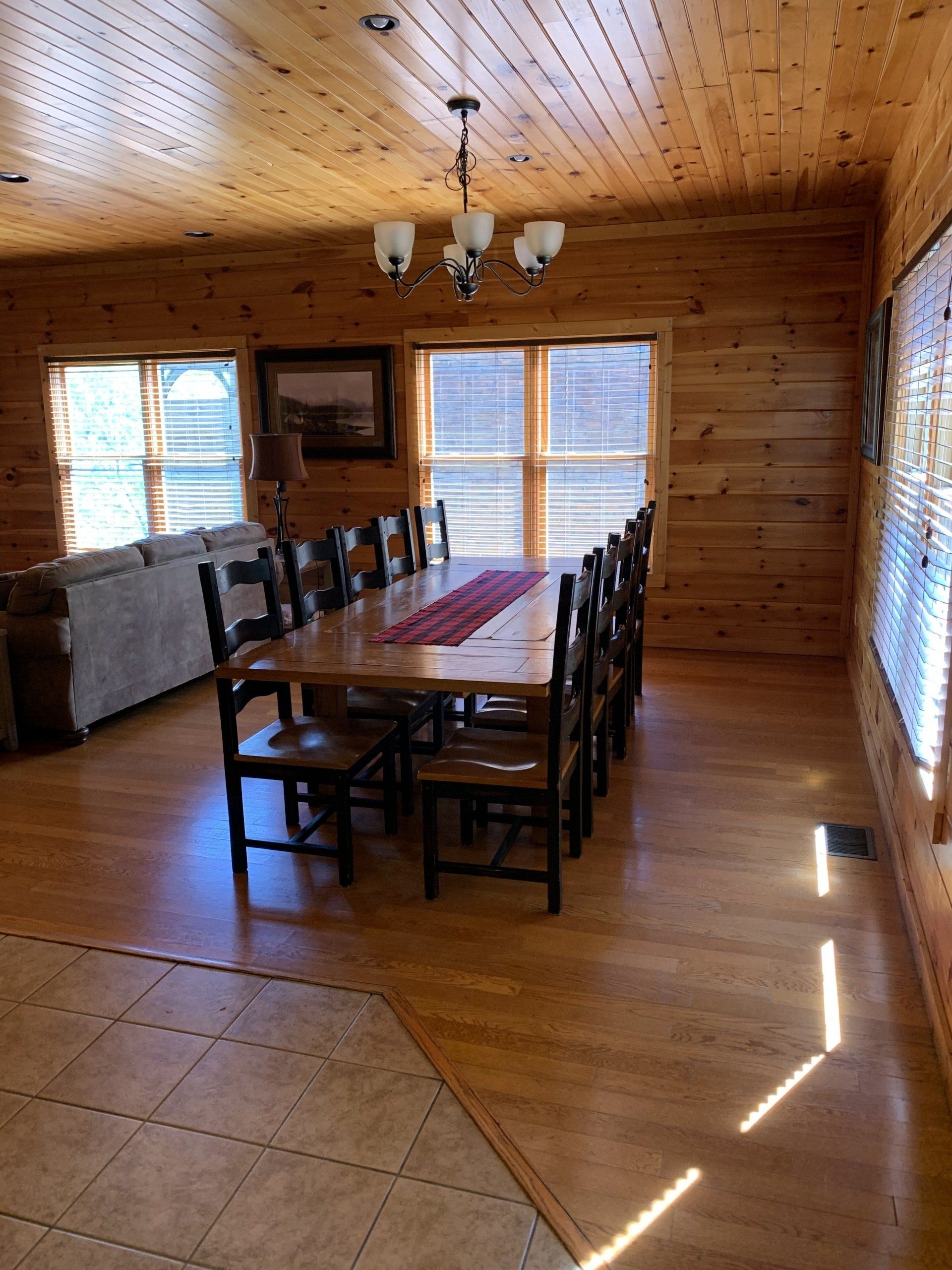 Wood-paneled dining room with a long table, chairs, tiled floor, and large windows letting in sunlight