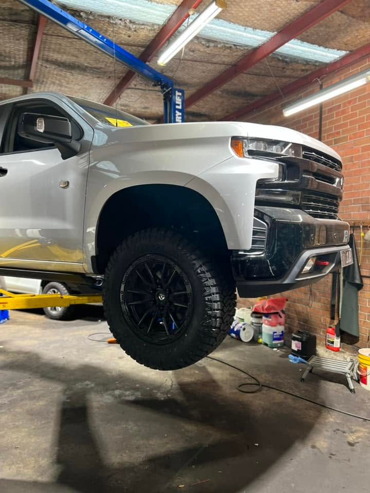 Silver pickup truck lifted on a jack in a garage, with black off-road wheels and tools nearby