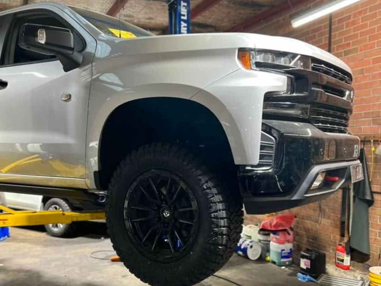 Silver pickup truck lifted on a shop lift inside a garage, with black wheels and off-road tires.