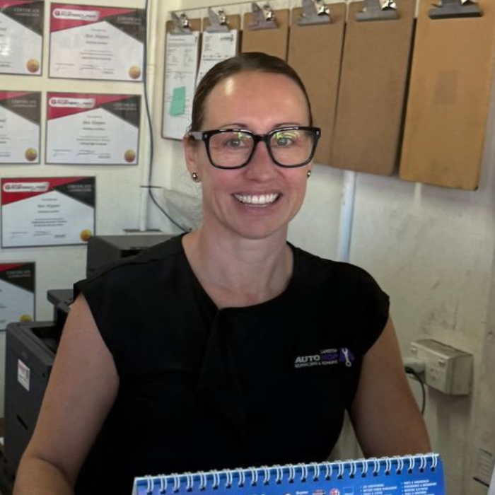 Smiling person in black top holding a blue clipboard in an office with certificates on the wall.
