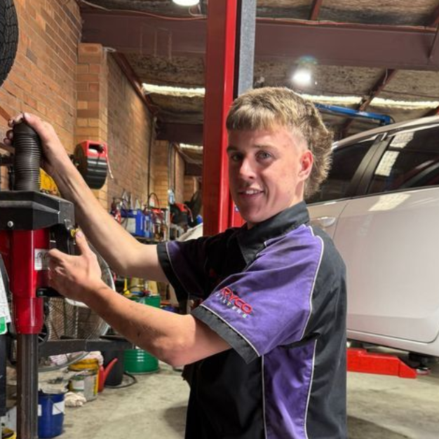 Mechanic smiling beside a white car in a workshop, adjusting a tool on a lift or machine.