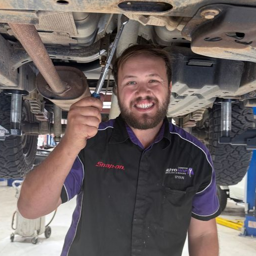 Mechanic smiling under a lifted vehicle, holding a wrench in an auto repair shop
