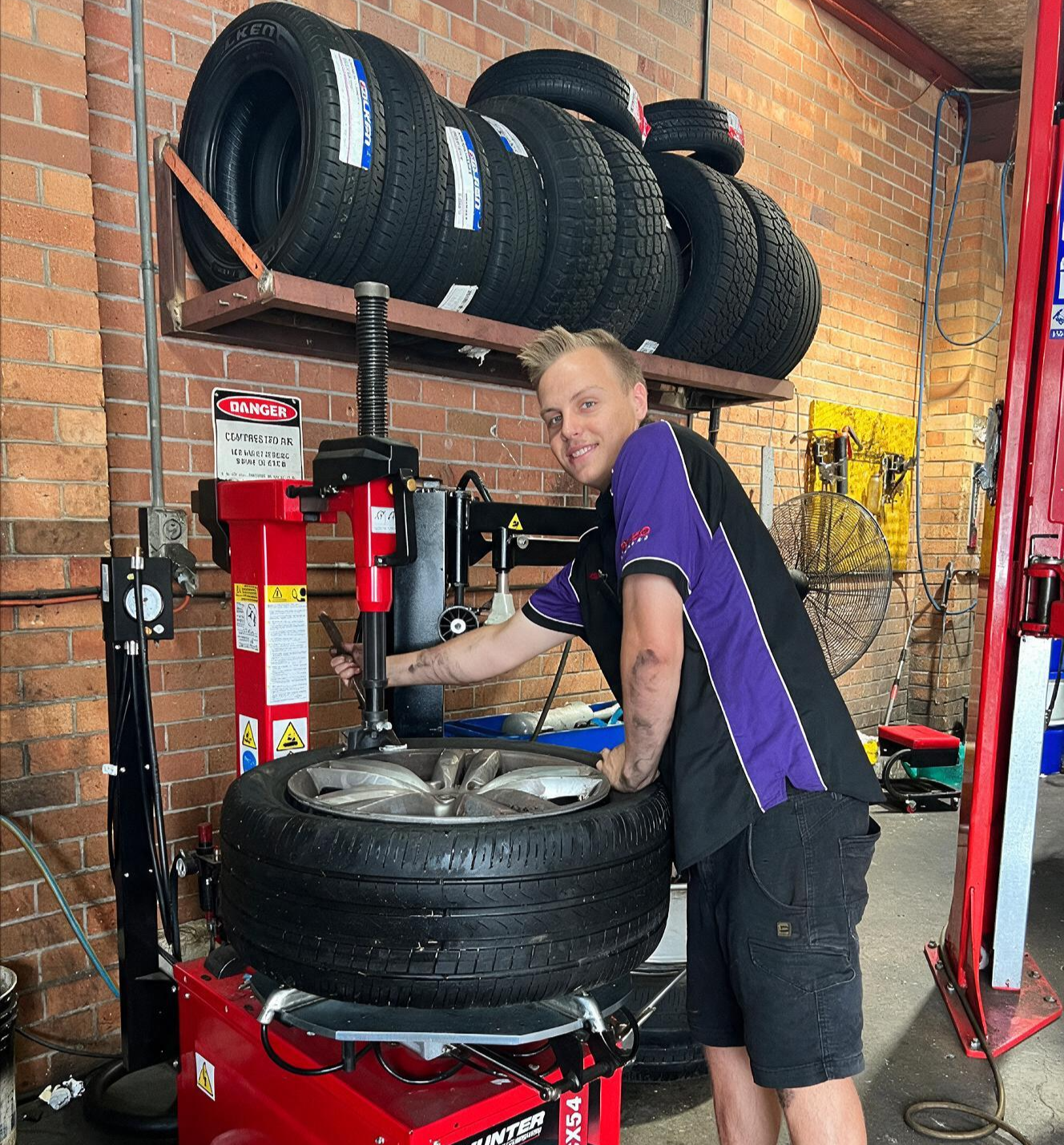 Mechanic in black and purple uniform changing a tire in an auto shop, with stacked tires overhead