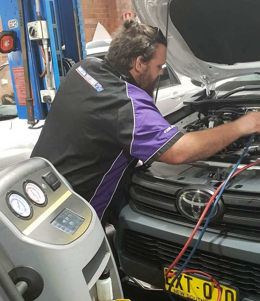 Mechanic in black and purple uniform working under a car hood in an auto repair shop.